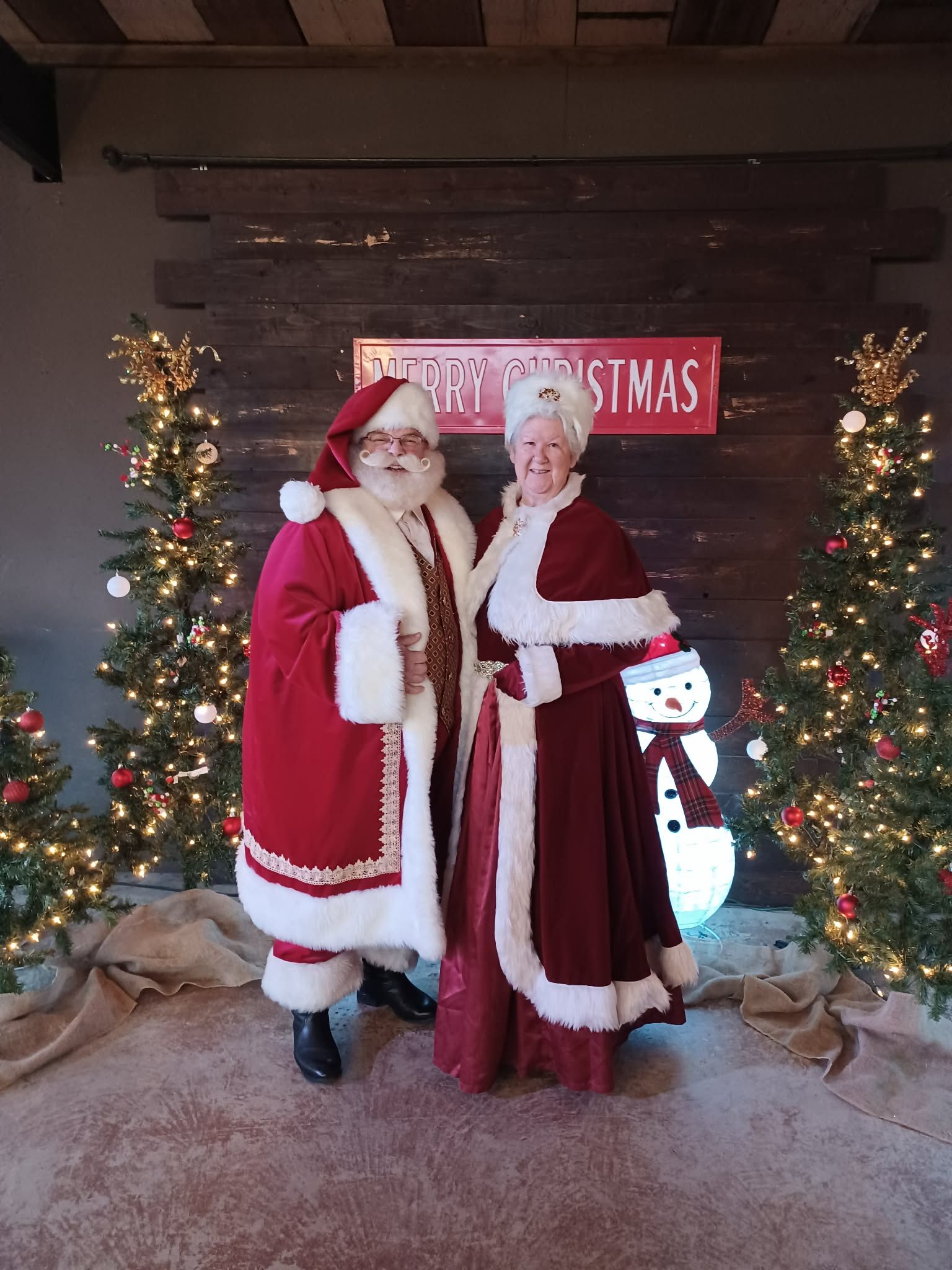 Santa and Mrs. Claus posed between lit Christmas trees beneath a Merry Christmas sign