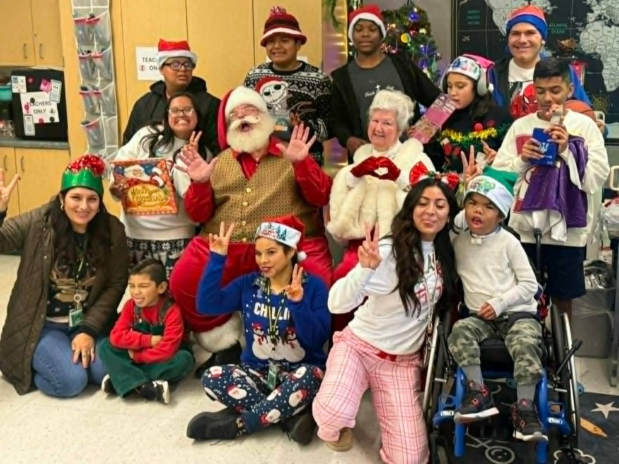 Santa and Mrs. Claus with students and staff in a decorated classroom, everyone waving at the camera